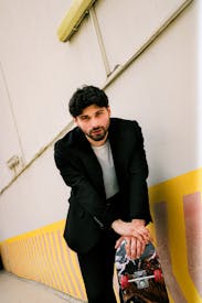 Man In Suit With Skateboard Leaning Against Wall